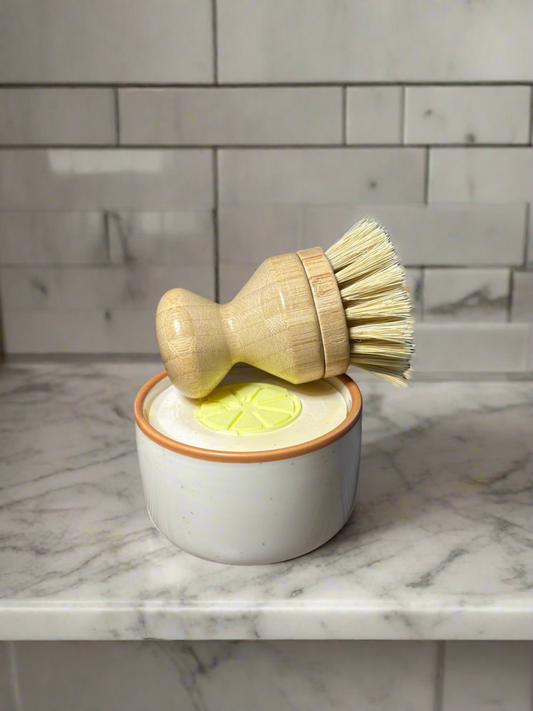 A beige ceramic ramekin with a brown trim on a marble surface, containing a solid dish soap. On top of the ramekin is a bamboo soft bristle pot scrubber with a removable head.